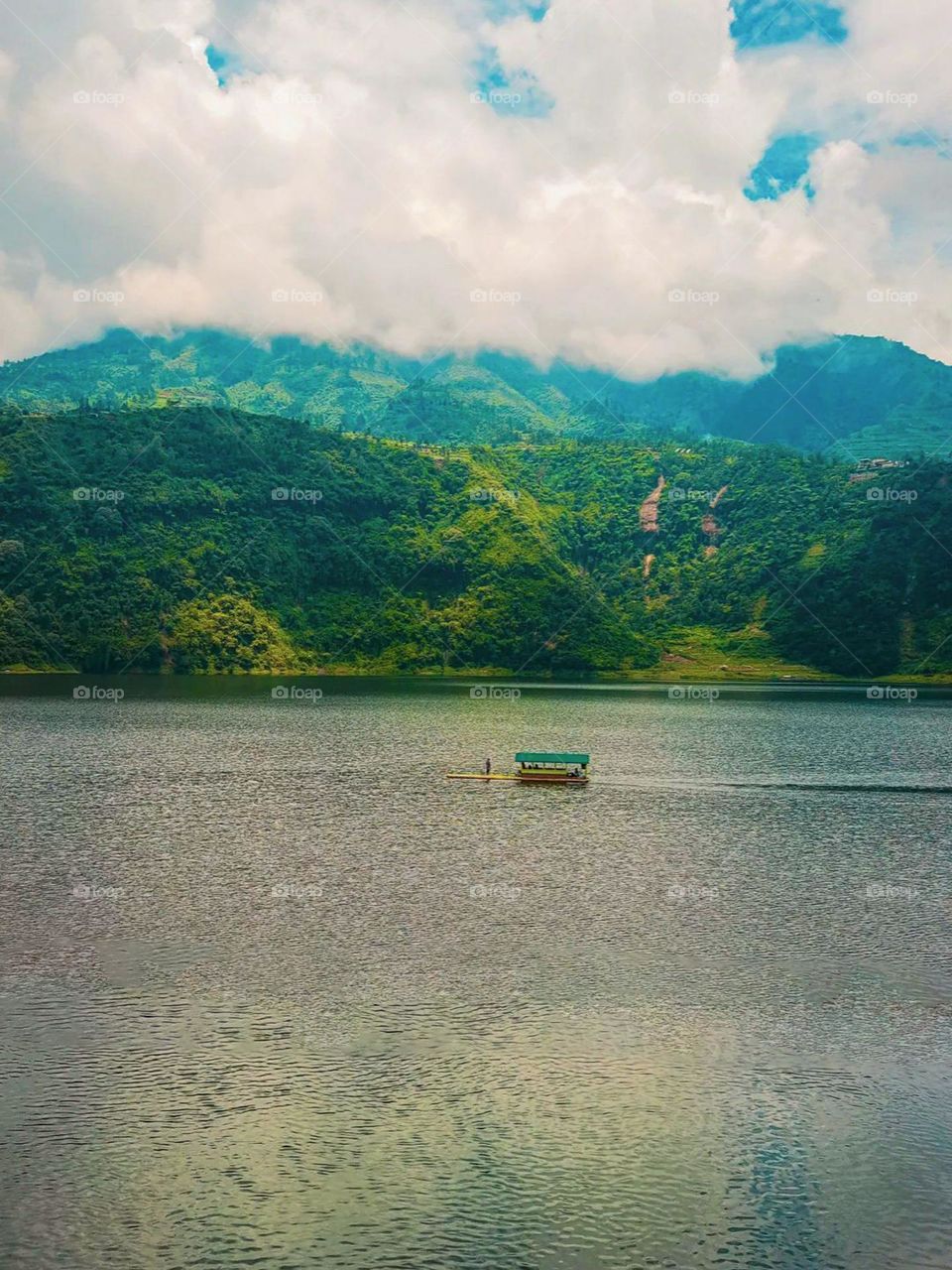 Long distance view of beautiful and calming natural scenery. This photo shows a small boat floating on a calm lake, with a backdrop of green mountains and white clouds