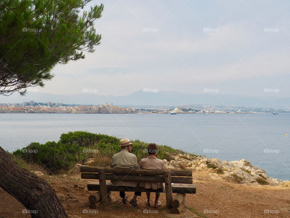 Elderly couple sitting on a bench enjoying the beautiful view of the sea off the coast of Antibes, France.