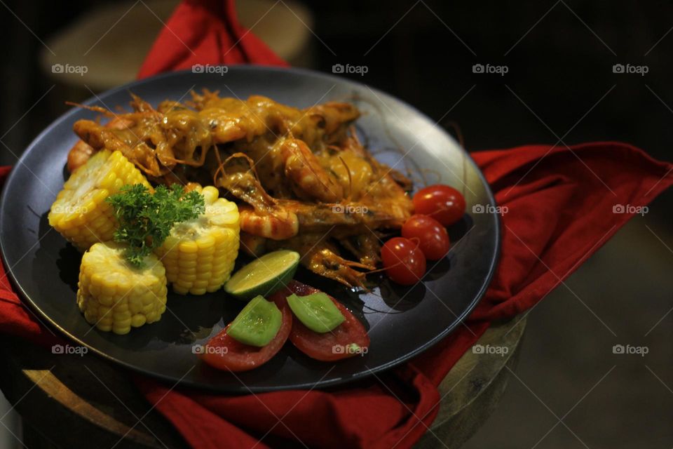 Asian food balado fried prawns with corn chunks, lemon slices and tomatoes served on black plate with red cloth bottom and wooden coaster. Asian style street seafood.