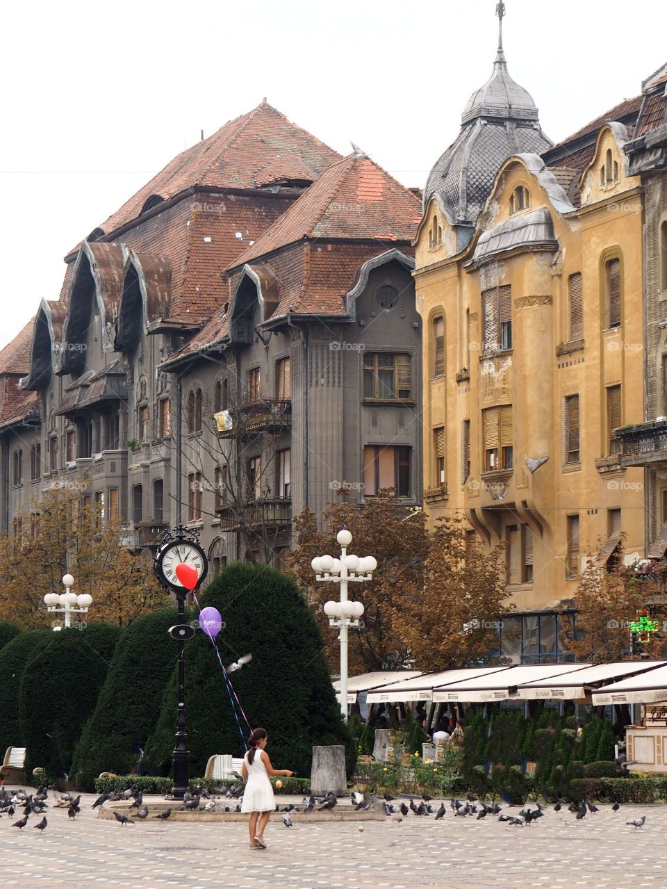 Little girl in a white dress with a red heart-shaped balloon standing on a square in the middle of the city surrounded by birds.