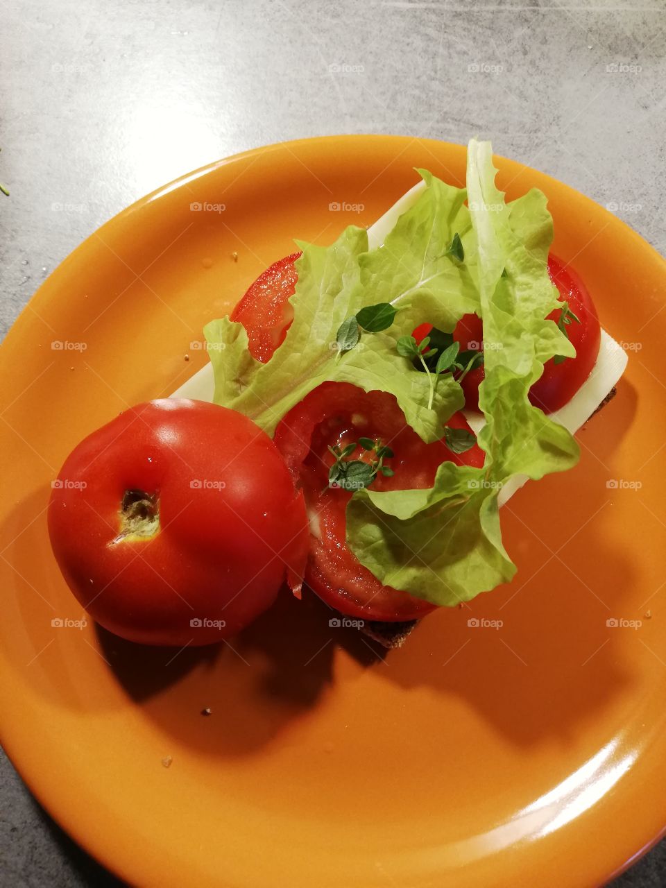 A slice of dark bread with butter, cheese, lettuce, tomato and some thyme on an orange plate on a grey table. Half of ripened tomato has the stem up. A water drop and some crumbs on the dish.