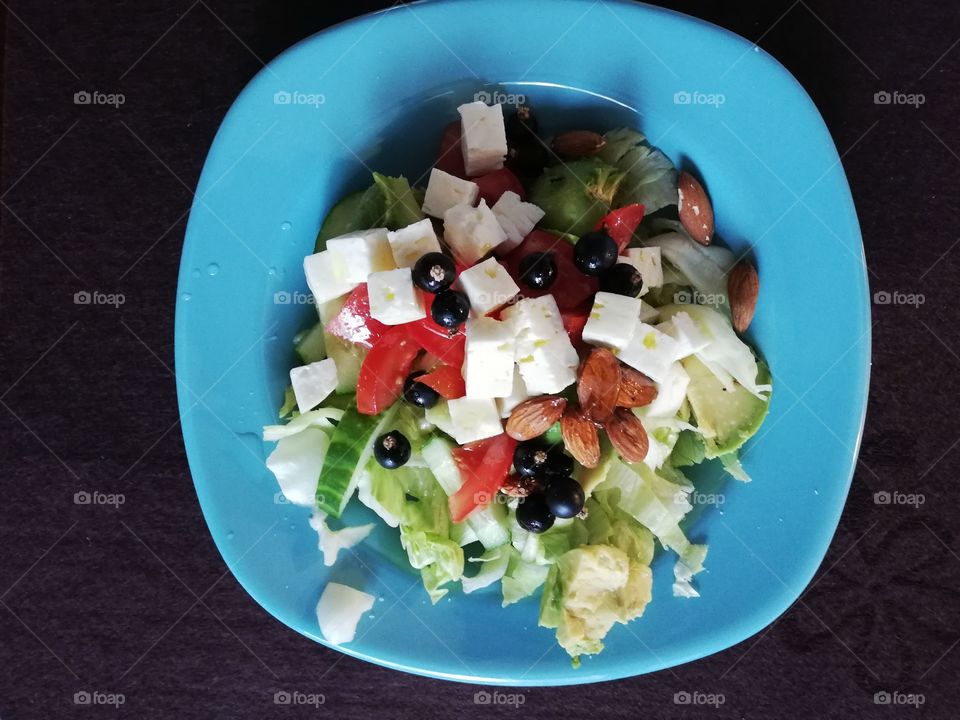 Blue plate is full of delicious vegetables and berries: tomato, cucumber, avocado, salad, black berries, cubes of cheese and almond with salad dressing.