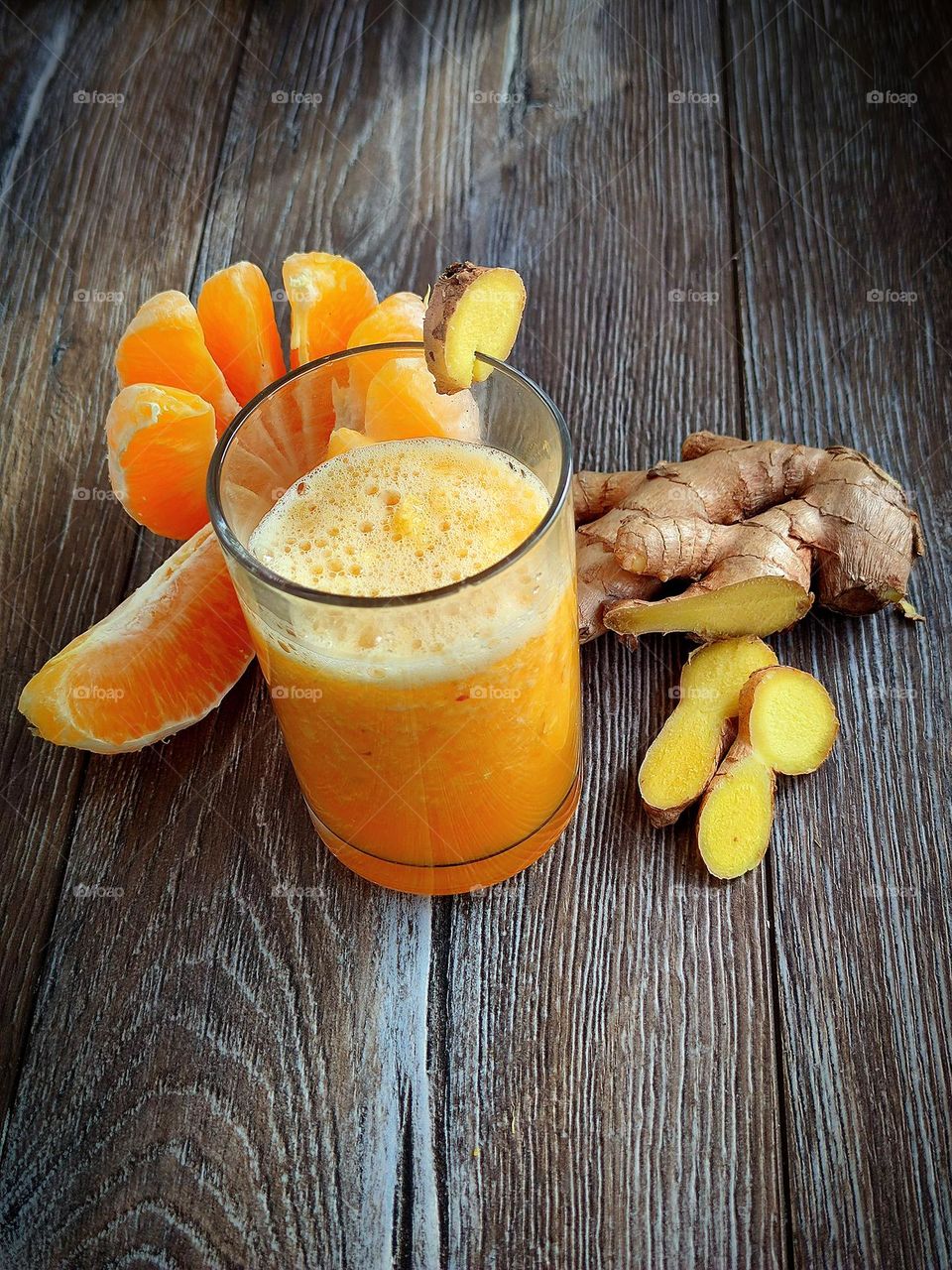 There is a glass with an orange smoothie on a wooden surface.  On the glass is a piece of ginger as a decoration.  Ingredients lie nearby: peeled orange, ginger root and chopped ginger