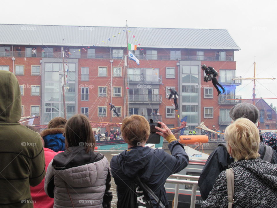 A crowd of people taking photos of men in jet suits hovering above Gloucester Docks and going at speed