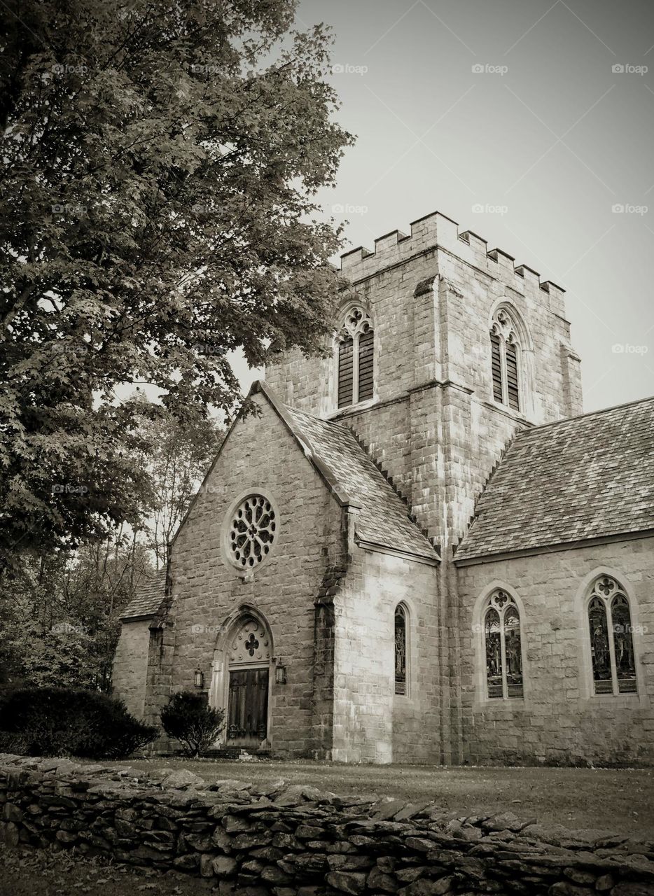 A black and white of a castle style old stone church building with a large maple tree outside in New England.