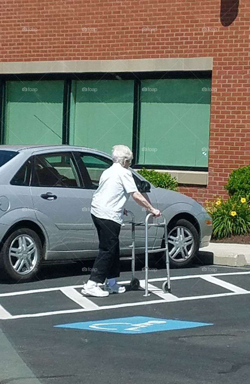 Older lady using walker, alone near car. Walking towards building.