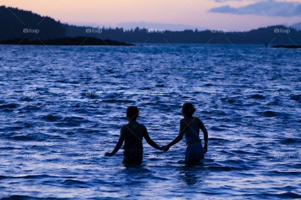 My 2 daughters, bravely inch hand-in-hand into the cold Pacific water to play in the last remnants of sunset.  The black silhouettes of their bodies & the coastal tree line are a stark contrast against the inky blue water and the pink & purple sky. 👥