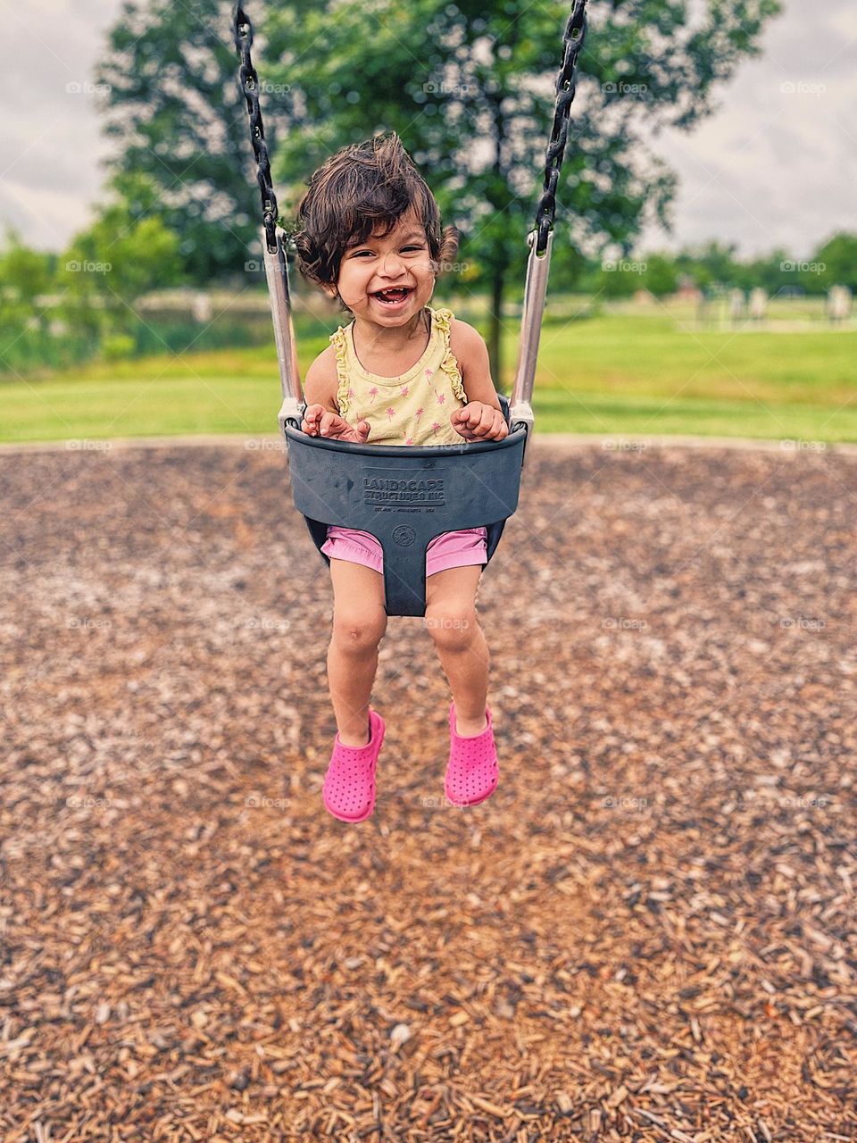 Toddler girl swinging at the park, toddler girl is happy on a swing, happy toddler at the park, toddler girl smiling from ear to ear, joyful toddler plays outside at the park, toddler girl portrait