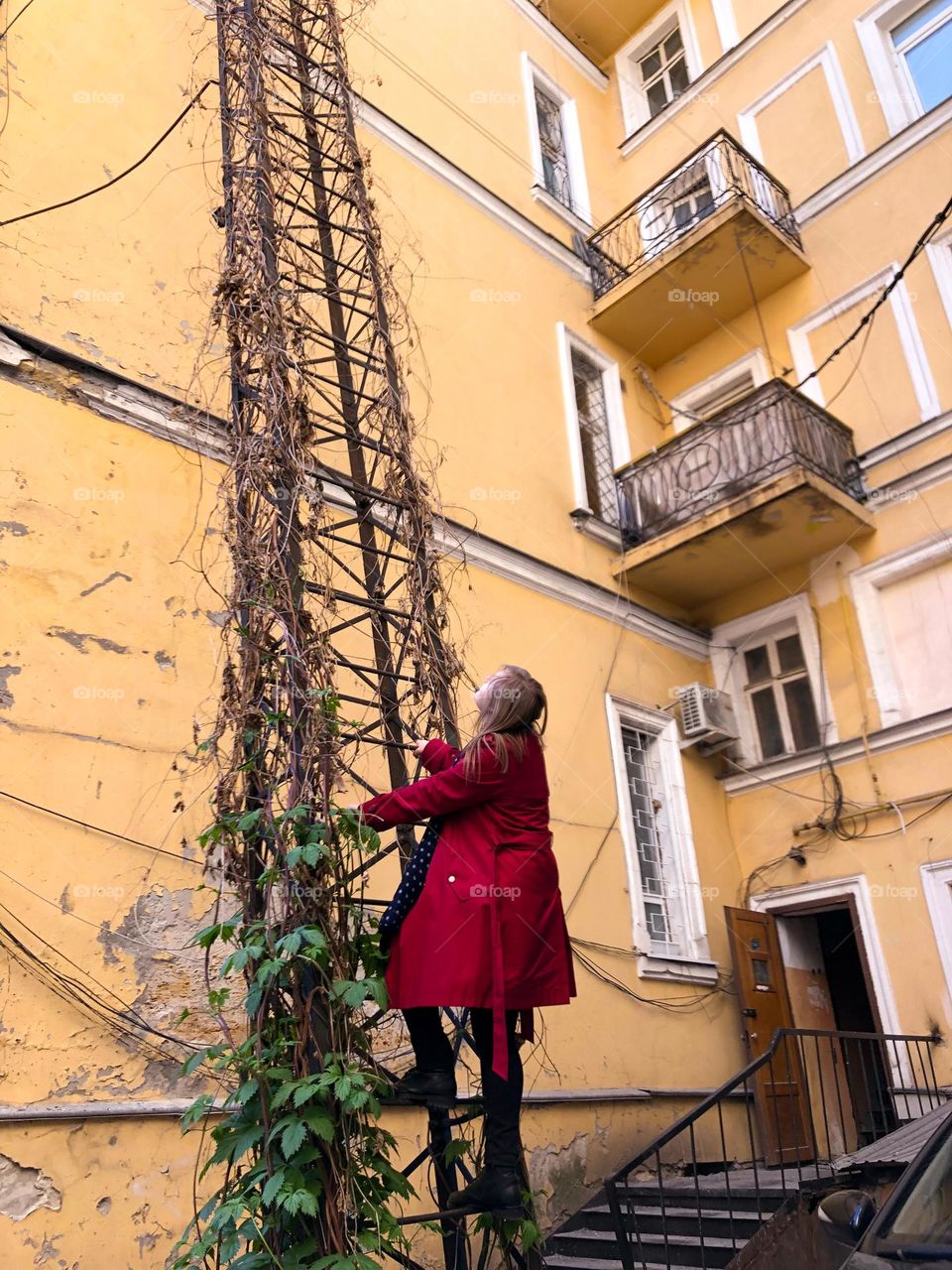 woman in red trench coat climbing ladder in old courtyard 