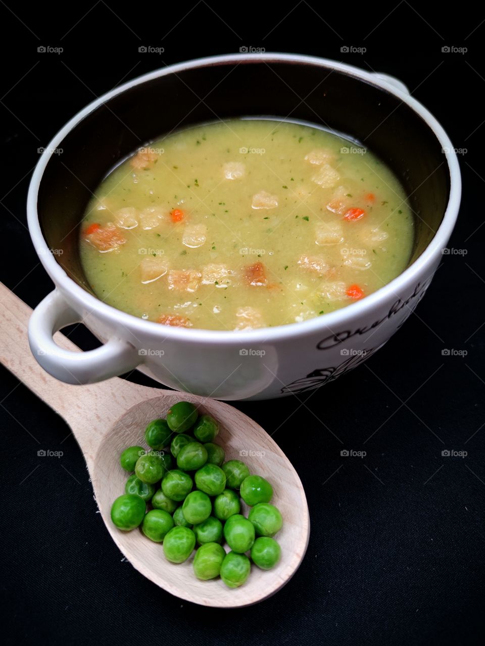 A deep bowl of puree pea soup and a wooden spoon with green peas next to it.  Black background
