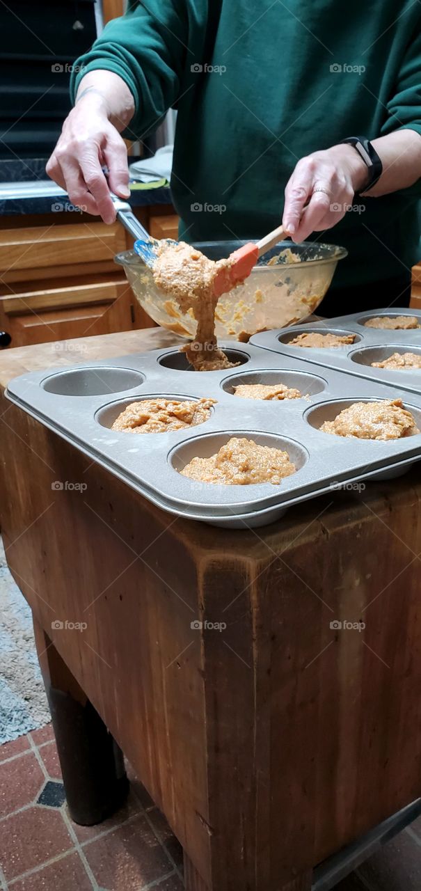 Filling muffin pans with homemade batter from glass bowl. Using spatulas to fill muffin cups.
