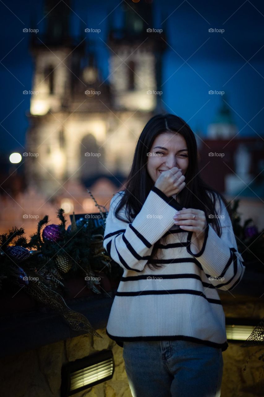 One young beautiful caucasian brunette girl with long hair laughs happily and embarrassed covering her eyes and mouth with her hand, stands on the balcony of a restaurant in Prague, close-up side view. Happy people concept.