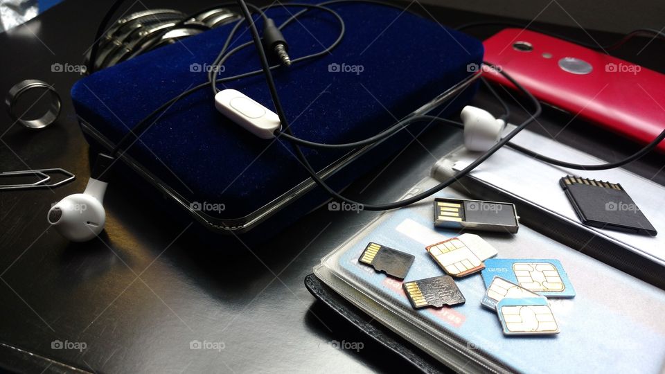 Mesa de escritório, com fone de ouvido, cartões de memória, chips de celulares, carteira, cartões de créditos, anel, relógio, clipe, smartphone...