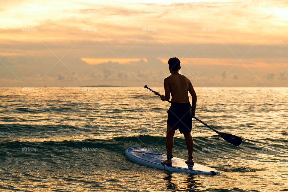 Slim men from behind using stand up board on the sea on sunset