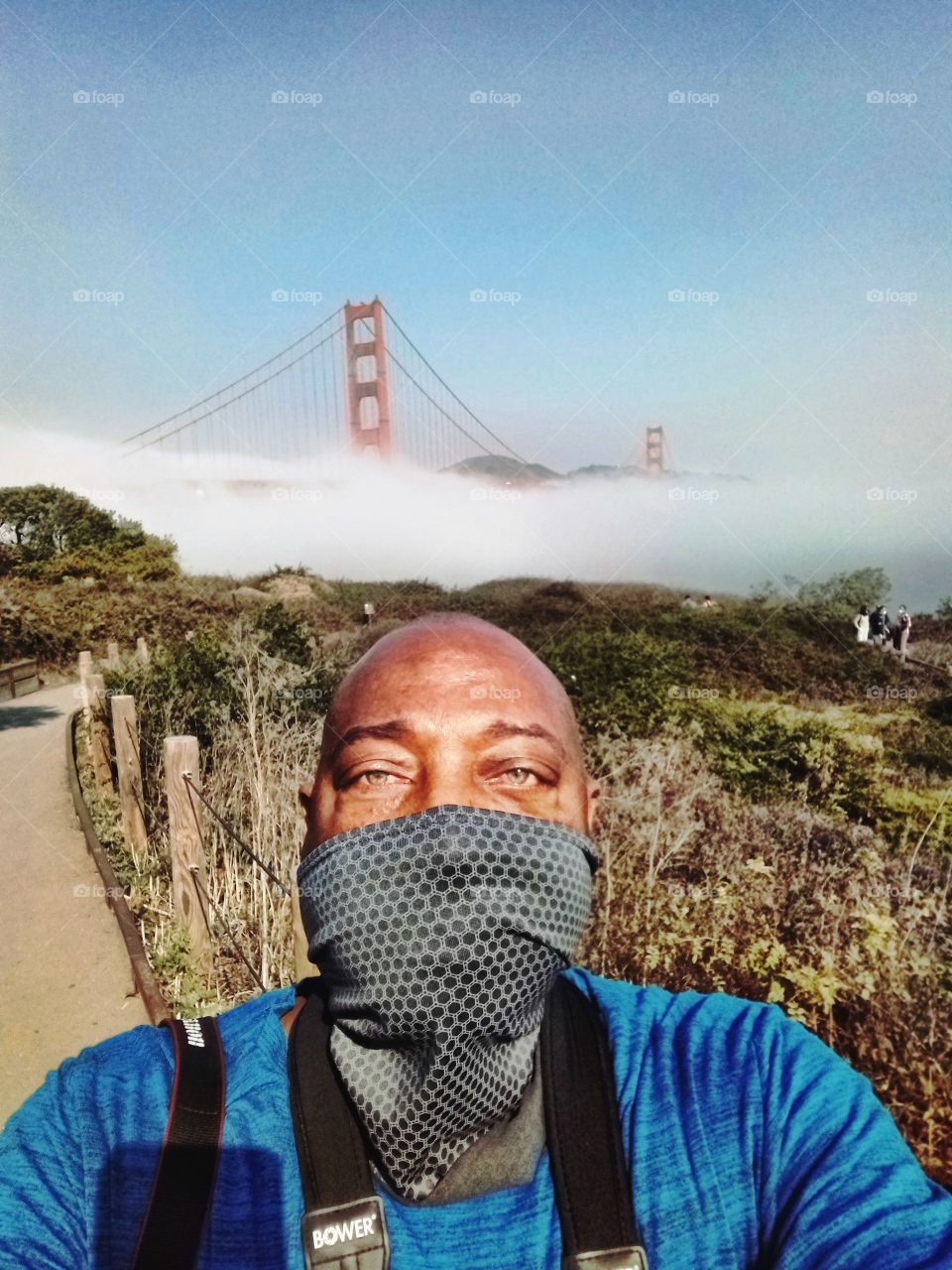 selfie, person wearing a face covering as the fog rolls in over the Golden gate bridge in the background.