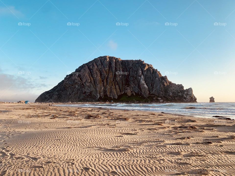 Morro Bay Rock and beach.  A gorgeous shorelines with blue skies and a soft sand beach with the trails or adventures all over.  The waves of the ocean crashing on the sand.  A beautiful landscape 