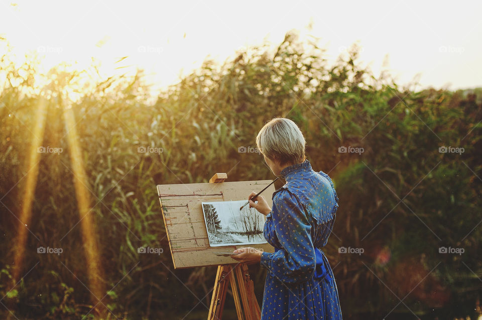 A woman artist in a long blue dress paints her picture in the fall by the river in orange sunset light.