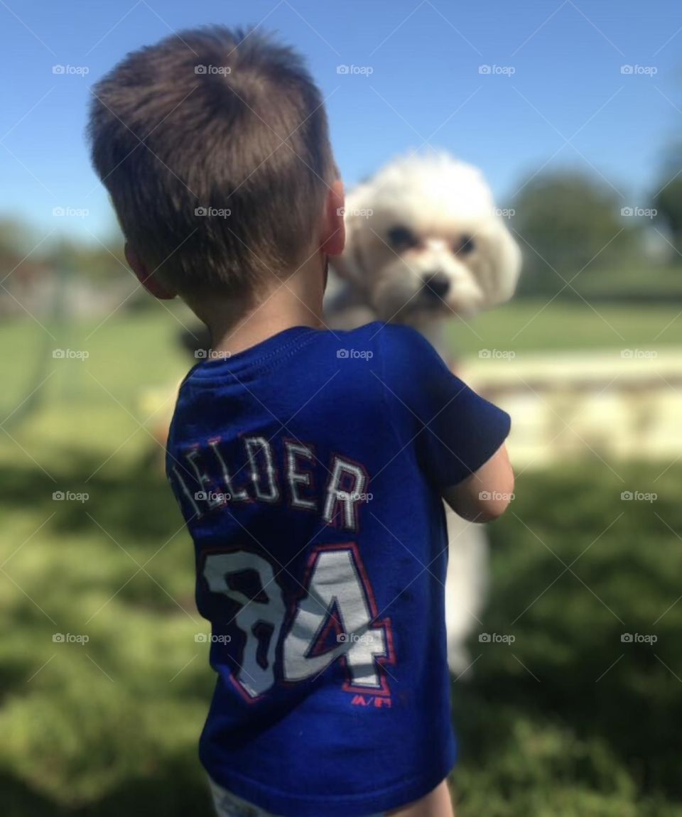 A young boy holds a white dog outside on the green grass. The sky is clear and blue with trees lining the background. 