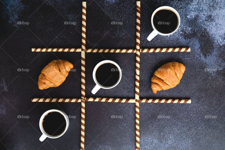 Business working morning with cup of hot coffee, sweet croissants on black background close up. Game of noughts and crosses. Top view, copy space, flat lay, mockup.