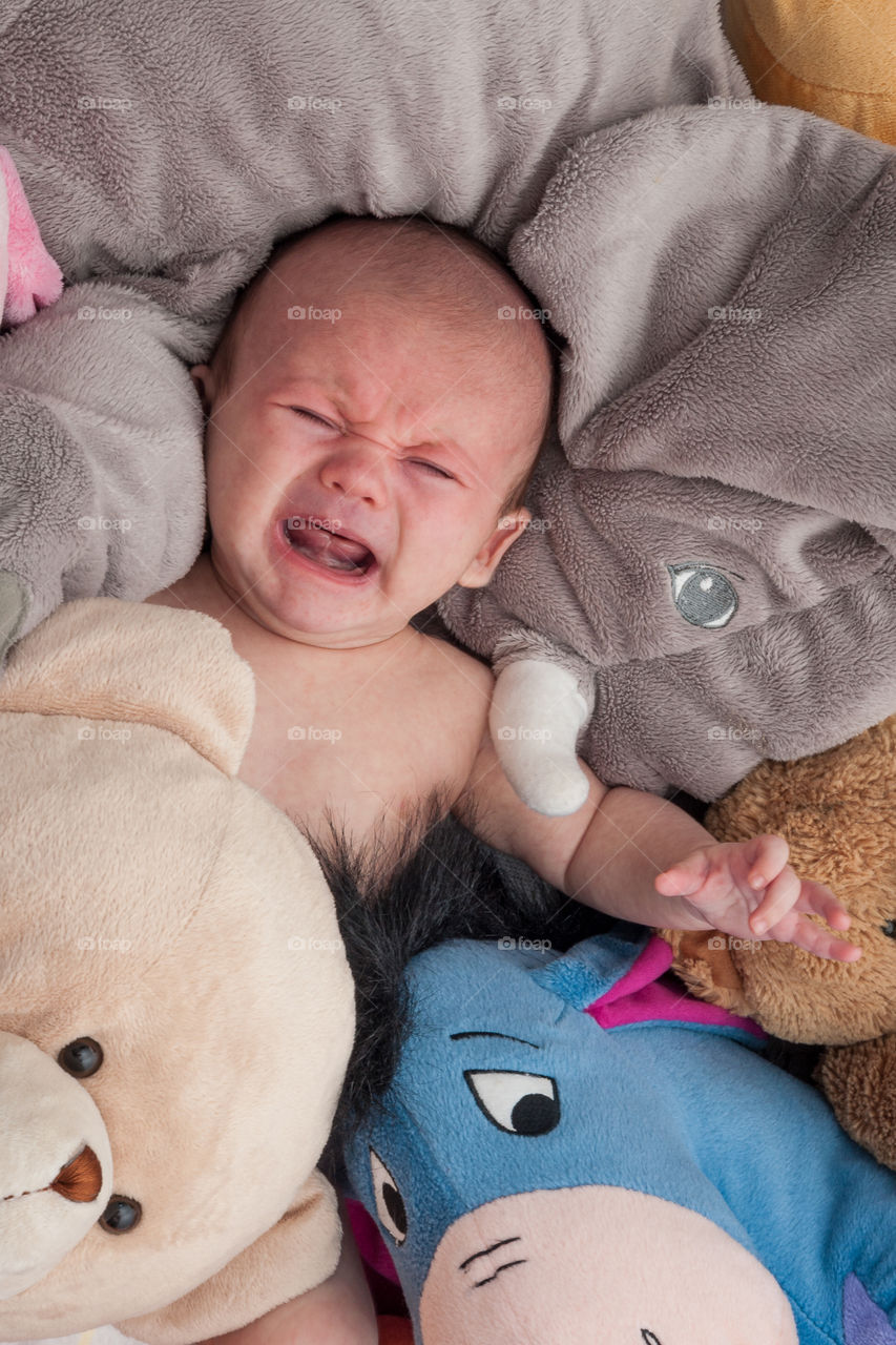 Infant in distress. Crying baby surrounded by soft toys, plush toys.