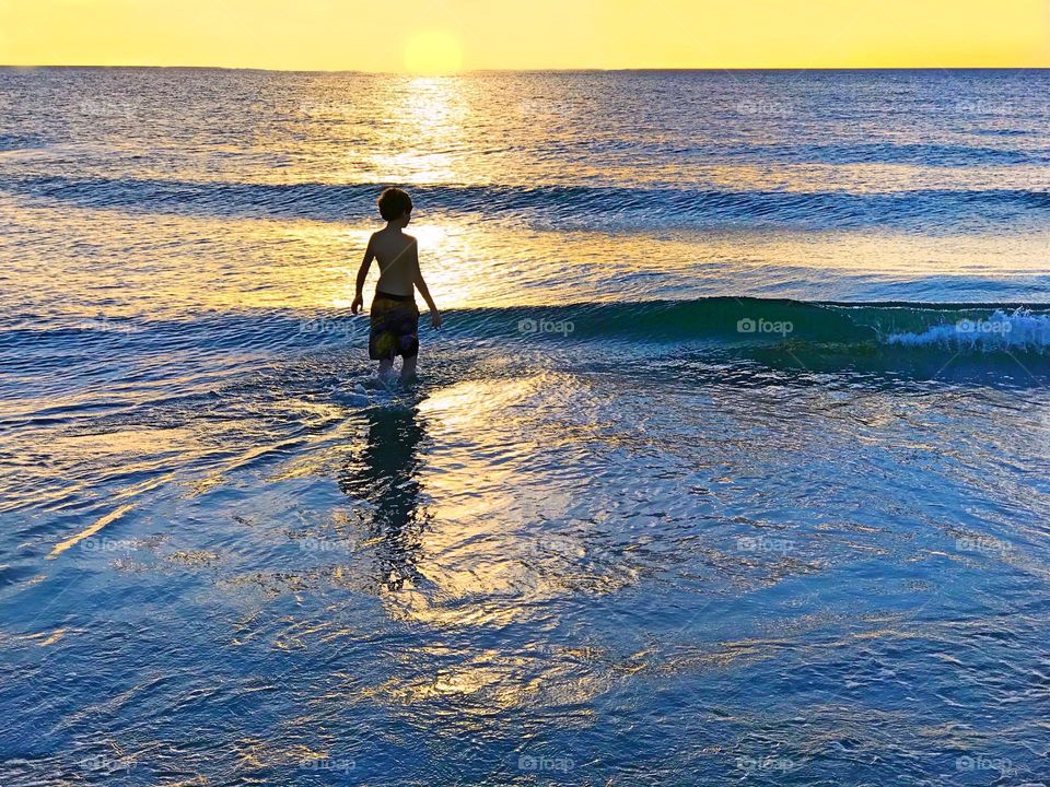 Small boy in the ocean surrounded by a bright yellow sunset.