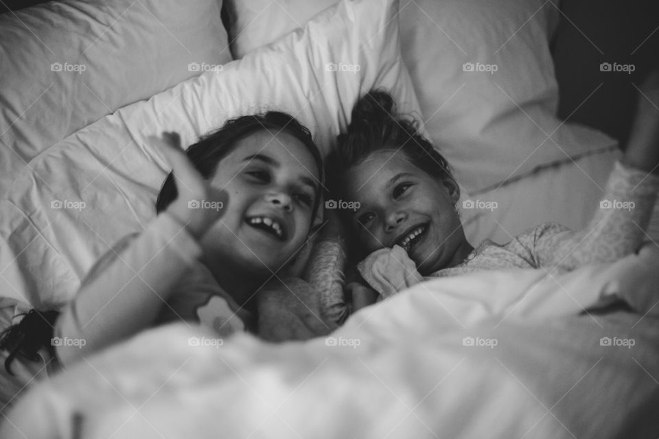 Black and white photo of two cute, young sisters in their pajamas, under the covers, laughing in bed together 