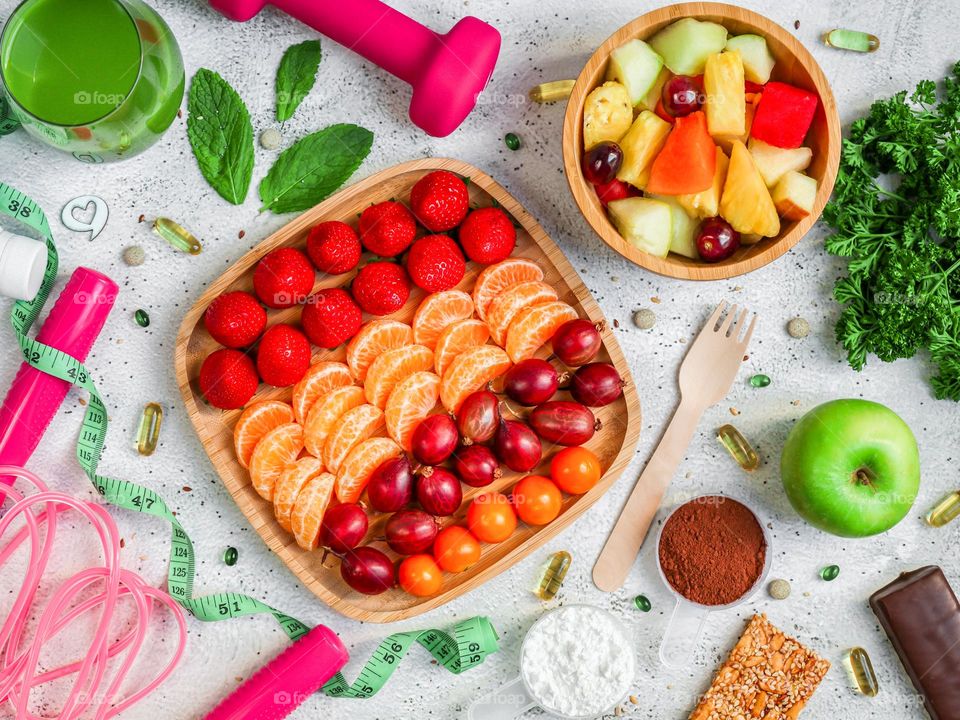 Fresh berries and fruits in wooden plates, dumbbells, measuring tape, seamstress, juice in a glass, bars and protein powder on a light stone background, flatlay close-up. Concept of healthy eating, healthy lifestyle.