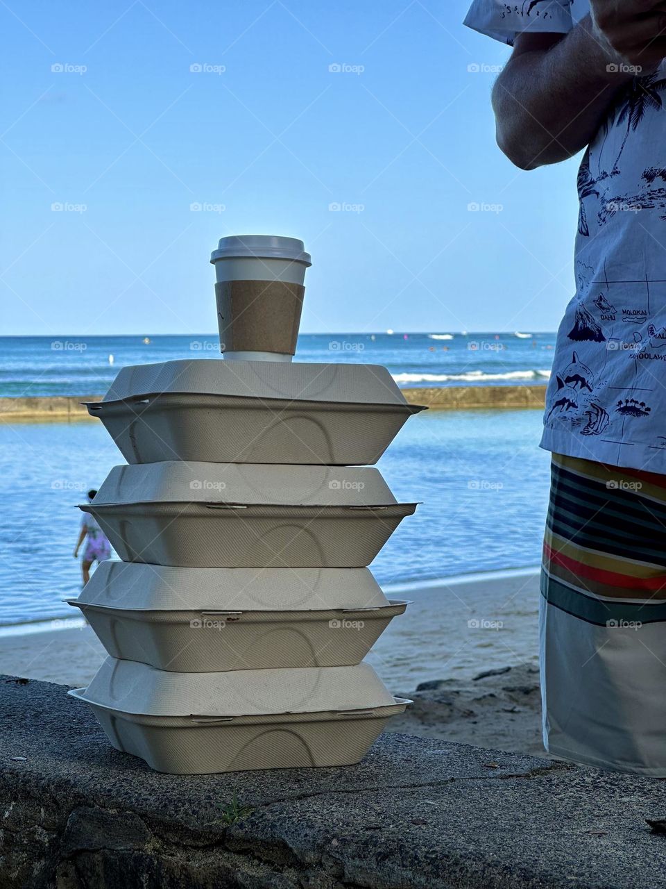 An early riser, in shorts and T-shirt, gets to bring breakfast for everybody on a vacation day at the beach. A visitor with a stack of to-go food orders in recyclable cardboard containers with a cup of hot coffee with a paper sleeve on top