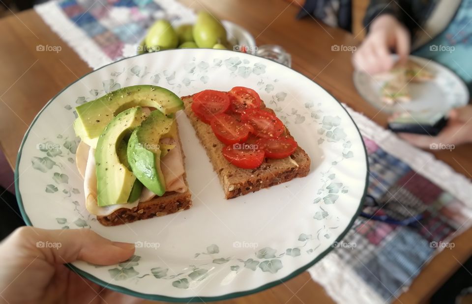 Avacado turkey tomato dijon mustard sandwich being placed on a table by a woman's hand with a man eating a sandwich sitting down at a table