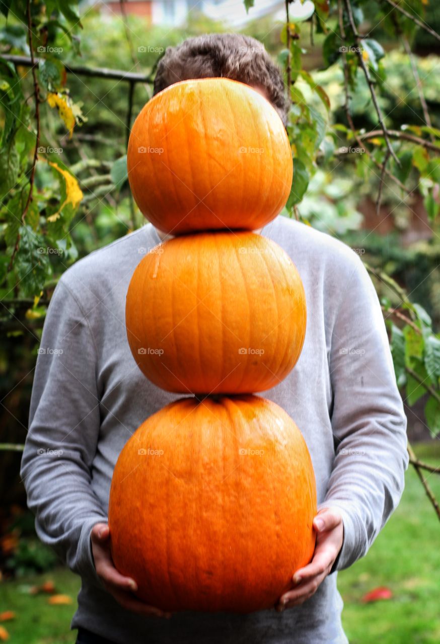 A young man standing in his garden and holding a stack of large pumpkins