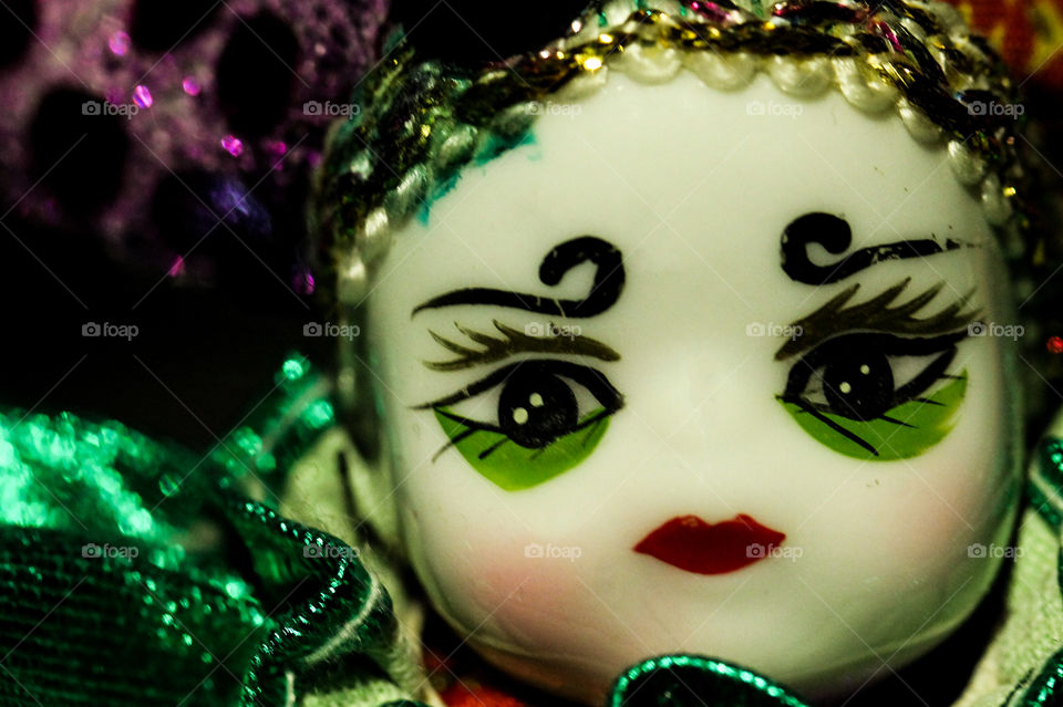 Macro closeup of the hand-painted face of a ceramic Venetian Jester doll. The doll is dressed in a colourful costume of purple, green, gold, red & blue with bells, sequins, ribbons and braided blonde hair. 