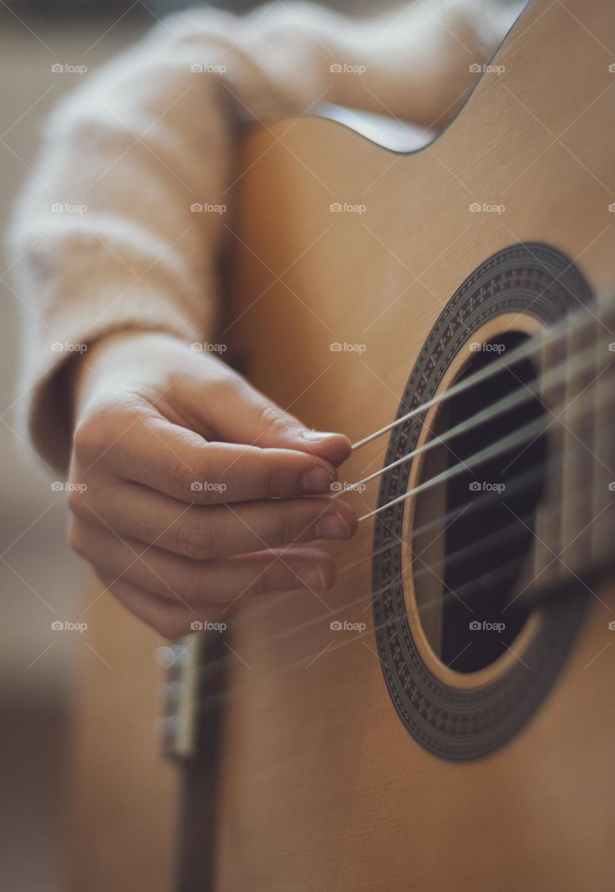 One hand of a little caucasian girl holds a guitar and picks the strings with her fingers while sitting on the sofa in the room in the morning and learns notes, close-up side view. Music education concept.