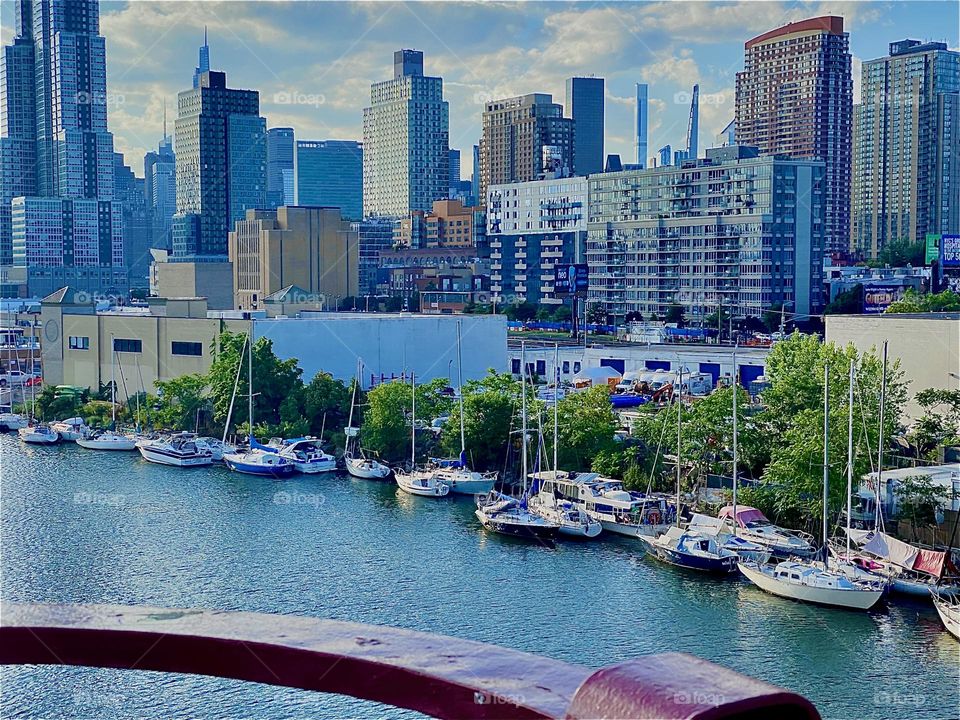 This is a beautiful view of “Newtown Creek” overlooking the “E River” from the “Pulaski Bridge”, the combined skylines of “LIC, Queens” & “Manhattan”. Motorboats & sailboats are lining the shore with its lush green trees. 2022. Hypnotic Productions