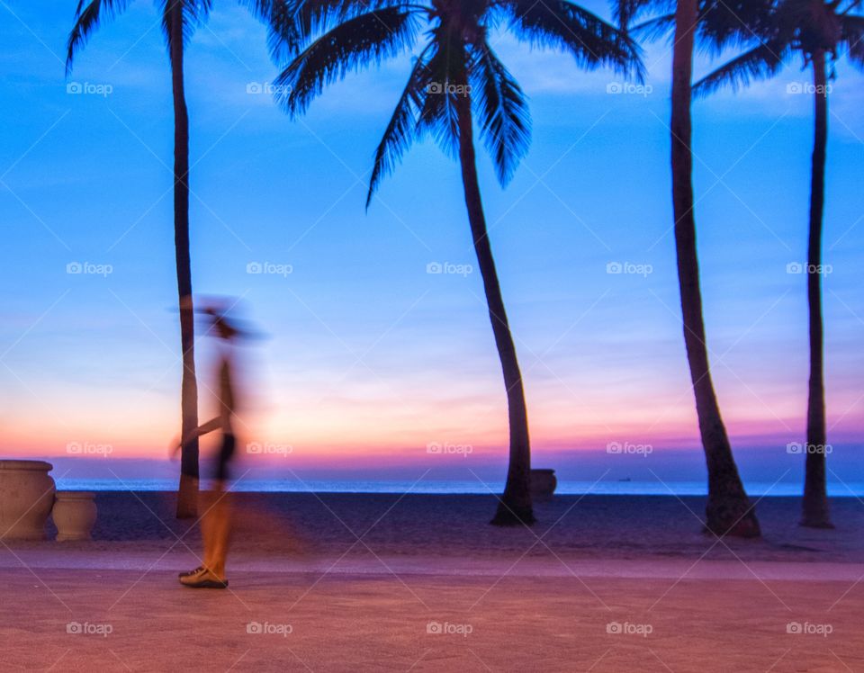 A walker is blurred as the sun creates silhouettes of the palm trees on an early morning sunrise at Hollywood Beach, Florida.  