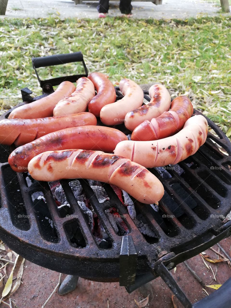 Two kind of sausages cooking on the black grill, made of iron. The ashes and charcoals are glowing red and the heat gives color and crispy surface on the shorties and open the cuts of the skin.