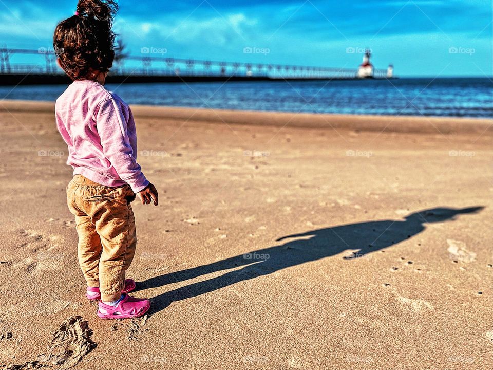 Beach landscape photo with toddler in foreground, beaches of Michigan, Michigan beach landscape photo, toddler looks at lighthouse, beaches and lighthouses, lighthouse in the distance, landscape photography by smartphone 