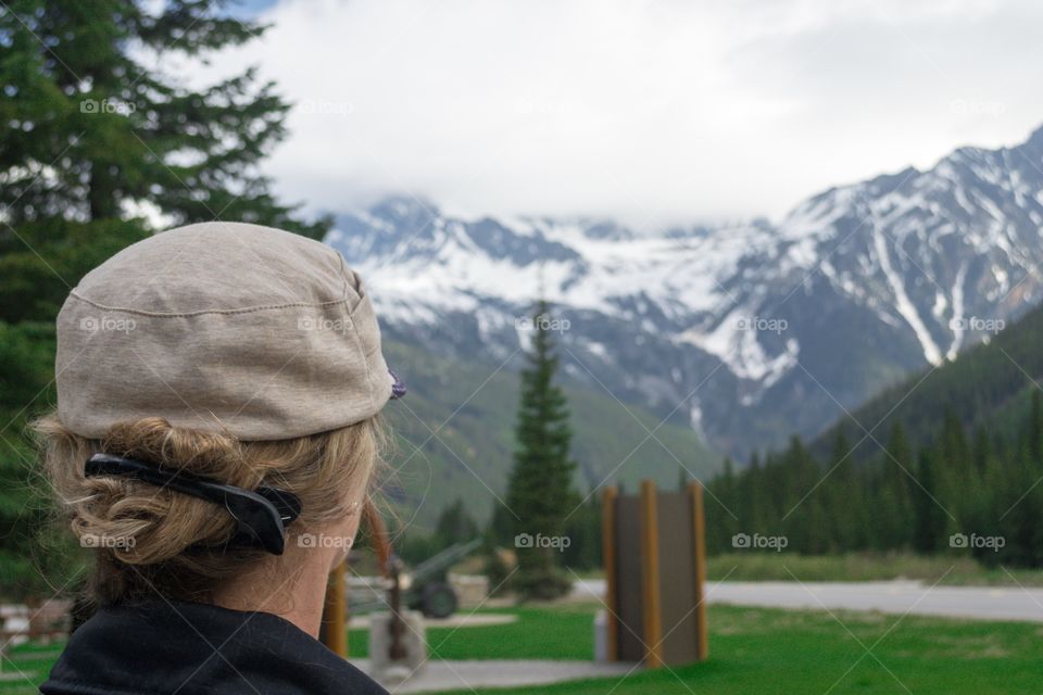 Woman (back of head) in hat foreground viewing spectacular scenery; Canada's Rocky Mountains, a snow alpine peak along the teams Canada highway in British Columbia at the border of Alberta outside glacier national park