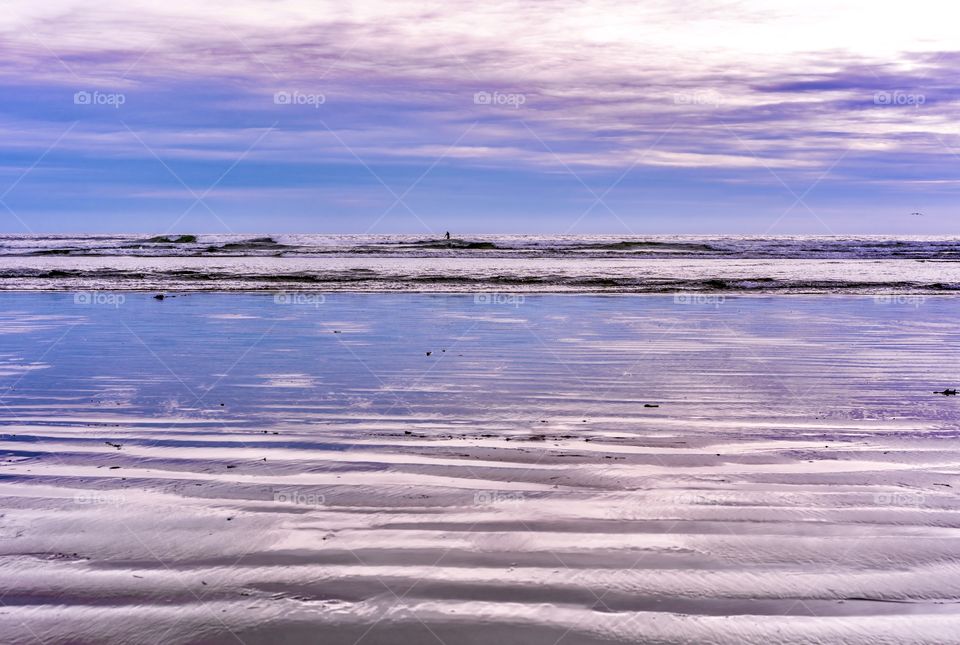 Surfer in a vast ocean scene wit purple sky, beach and ocean waves 