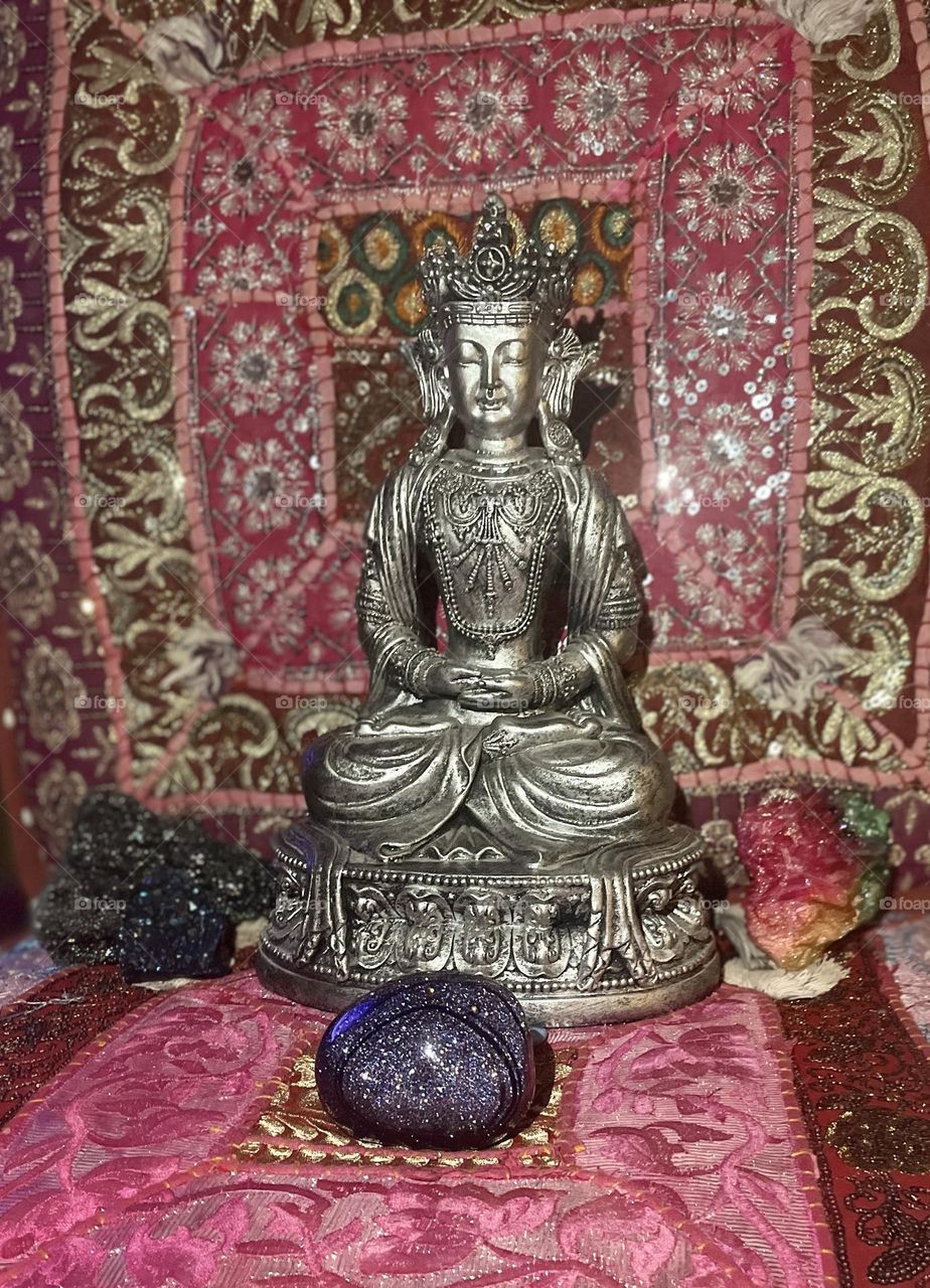 A silver Buddha statue meditating amongst several beautiful and colorful crystals with a colorful background.