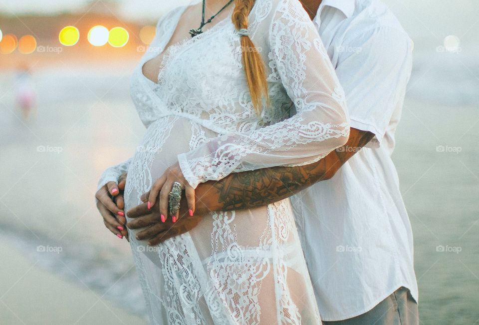 Tender and sensual photo on the beach. White dress emphasize the belly, husband hands show the love. Best photosession for our son 