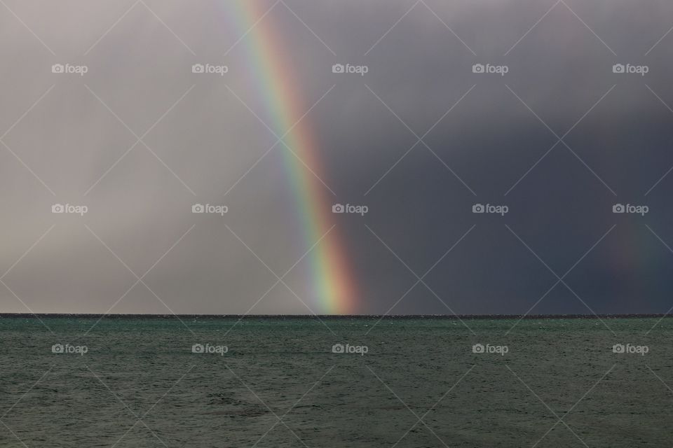 Dark, rain clouds brightened by colourful rainbow on ocean horizon 