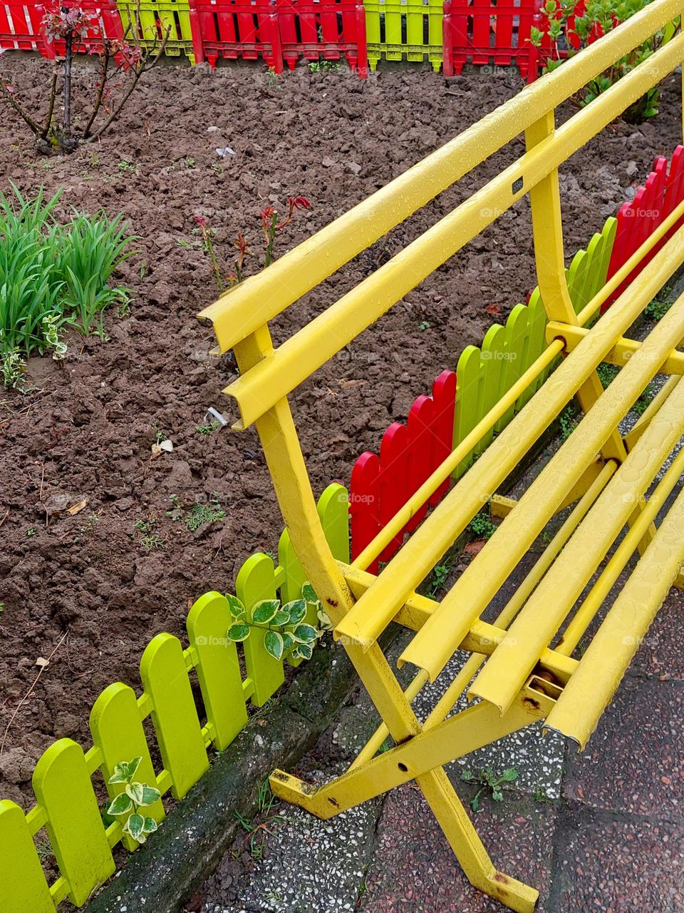 A yellow bench next to a garden surrounded by a red and green fence.  Dominance geometric shapes: rectangle and triangle