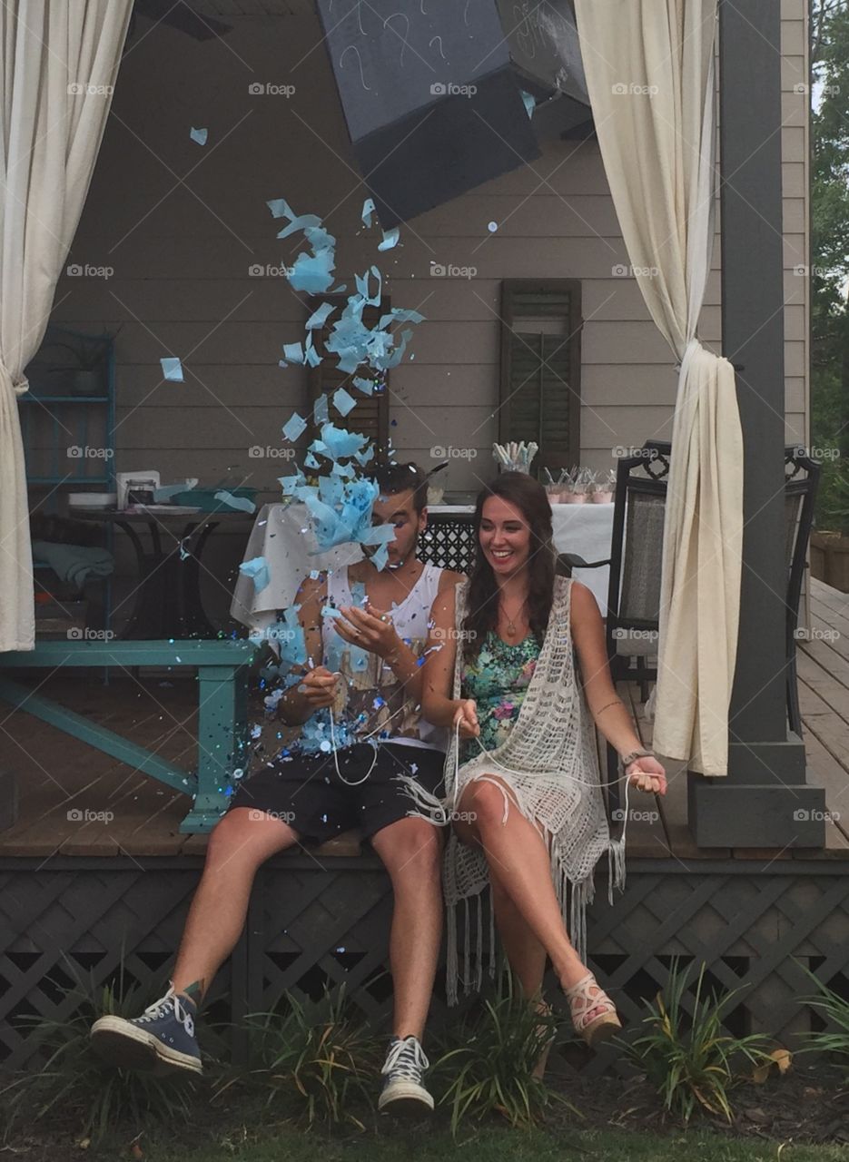 Gender reveal party with young couple finding out there baby is a boy with blue confetti paper and balloons falling.