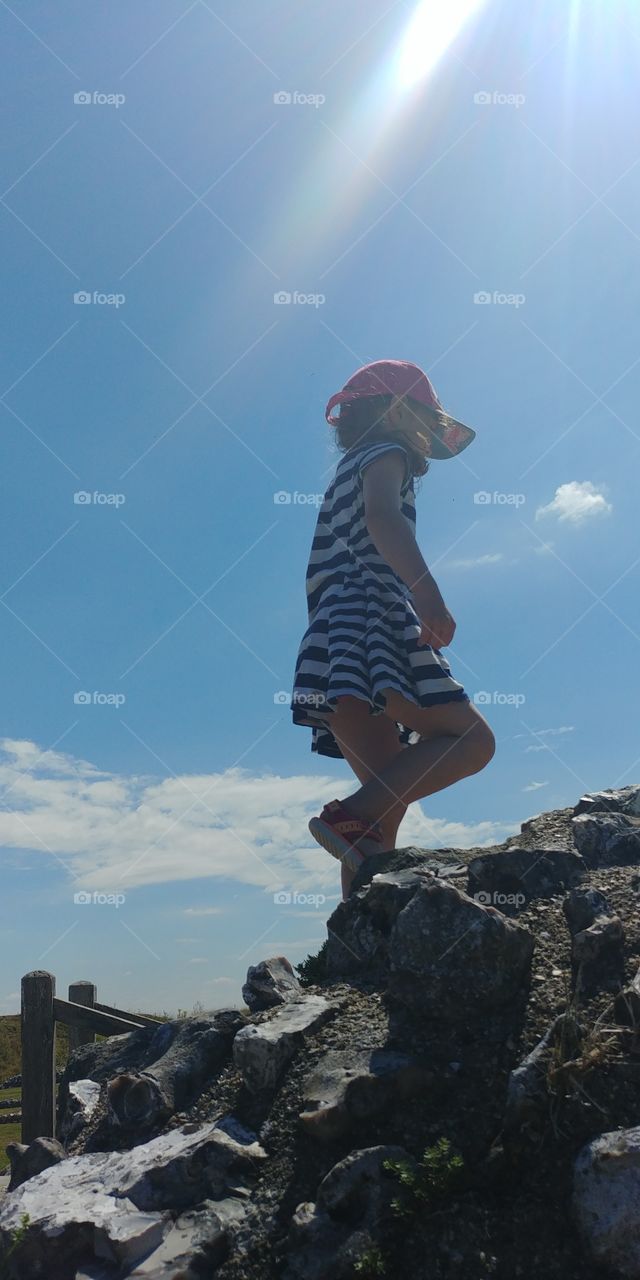 little girl in dress and cap walking up rocks in summer against blue sky.