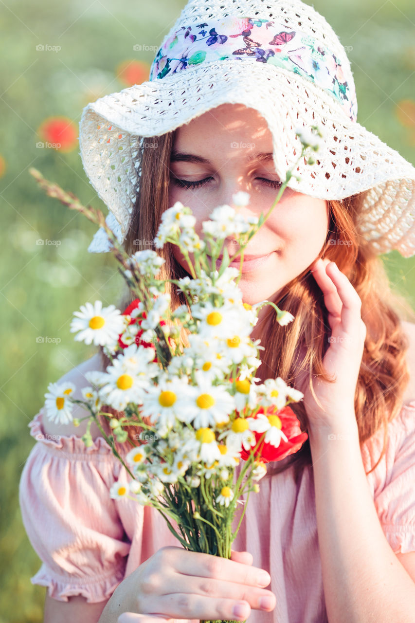 Beautieful young girl in the field of wild flowers. Teenage girl picking the spring flowers in the meadow, holding bouquet of flowers. She wearing hat and summer clothes. Spending time close to nature