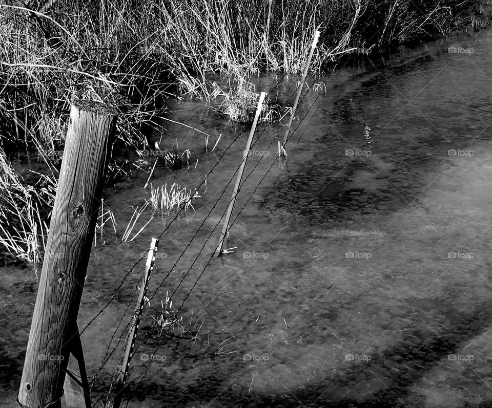 A barbed wire fence with wooden and metal posts partially under water in a frozen pond in rural Central Oregon on a winter day. 
