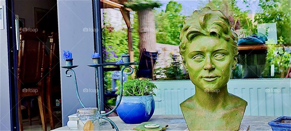 A beautiful bronze bust has been taken out onto the terrace in “Bavaria”, Germany where it sits on a table next to a potted plant. The lush green garden reflects in the large glass sliding door and facade of the bungalow. 2024. Hypnotic Productions
