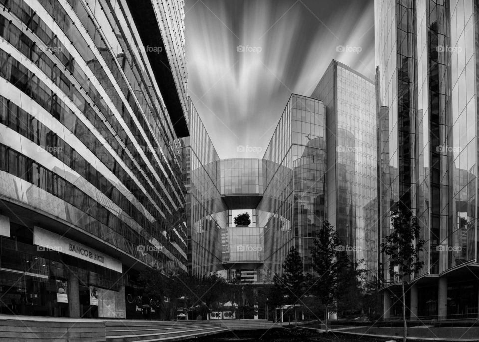 black and white photograph of a set of buildings with a small park in the center and leaking clouds