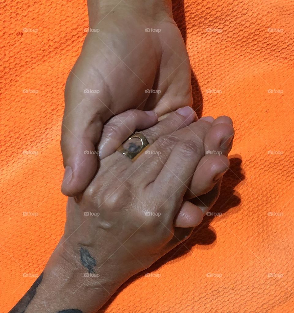 A couple holding hands in a very tender and caring way over an orange bed sheet. 