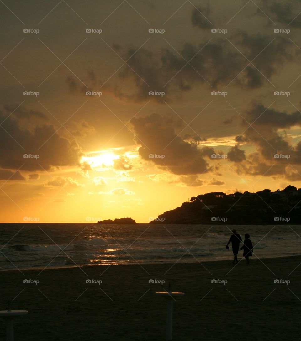 Husband and wife take a walk on beach while sun shines among clouds in orange light.